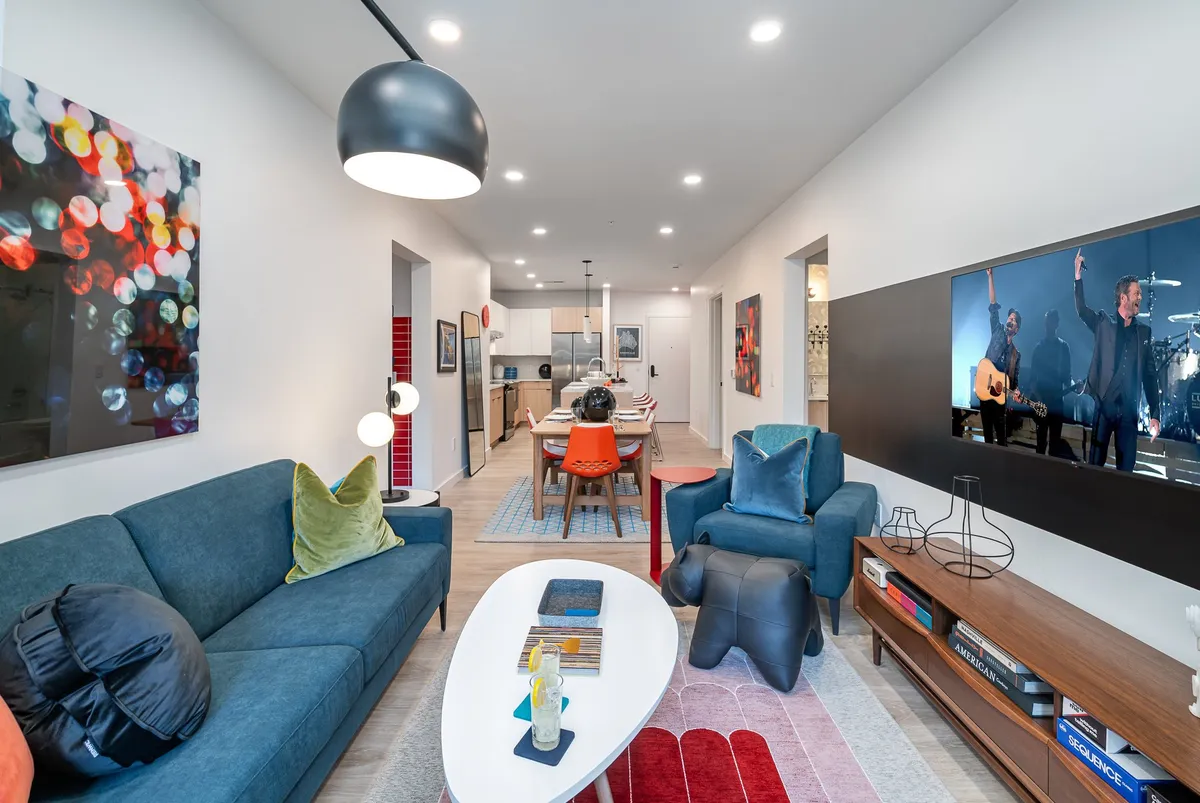 The image showcases a vibrant and stylish living and dining area featuring a plush blue sofa, colorful modern wall art, a sleek dining table set for entertaining, and a TV displaying a live music performance. The open layout invites conversation and relaxation, echoing Nashville's lively spirit.