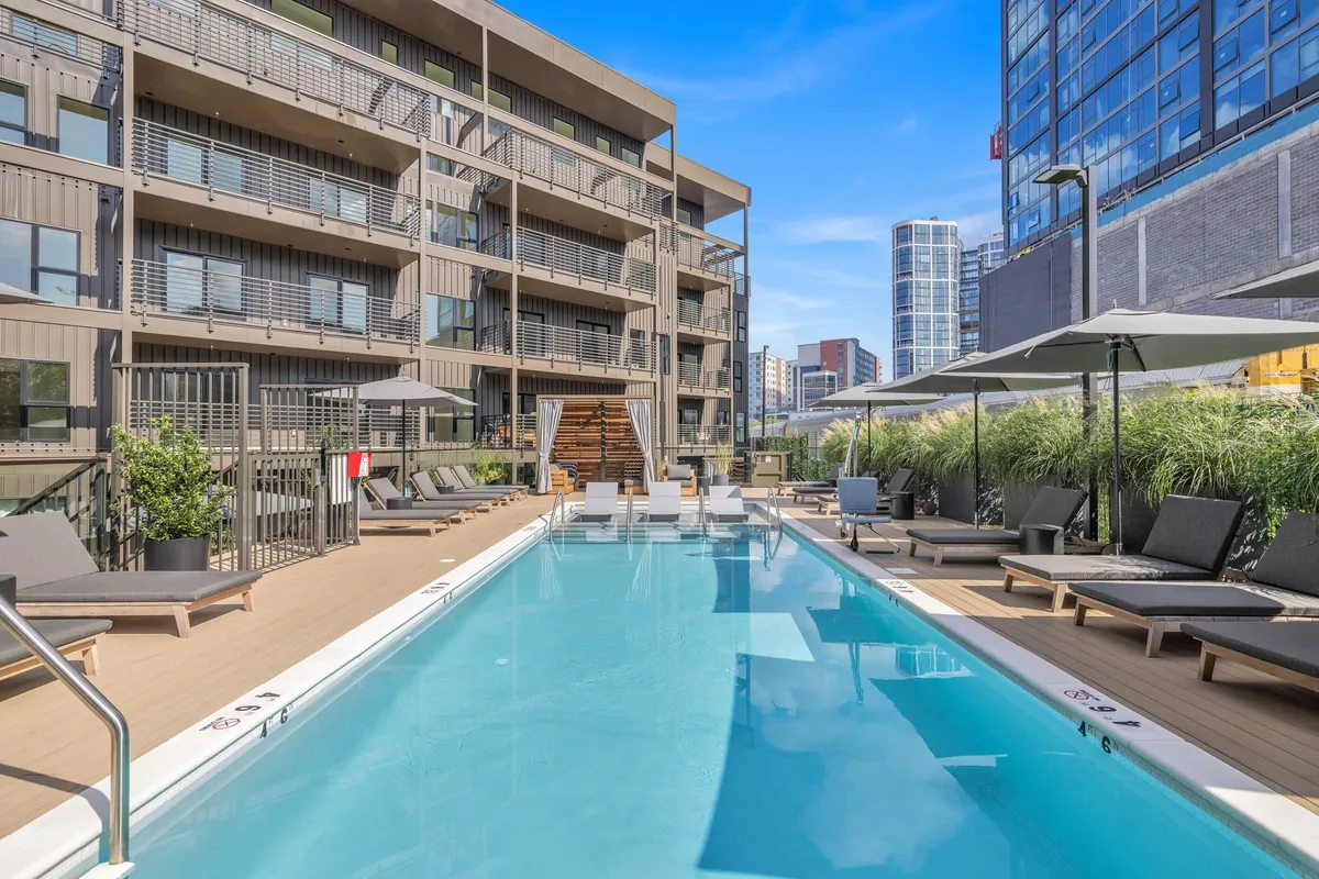 A resort-style swimming pool is surrounded by sleek lounge chairs and lush greenery, with the building's modern facade in the background. The area exudes a relaxed, social atmosphere ideal for warm-weather enjoyment, poolside gatherings, and sunbathing.