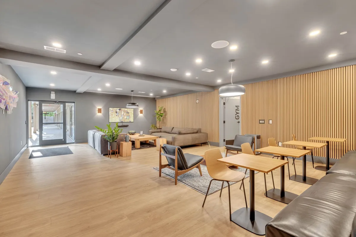 A spacious building common area with lounge seating, dining tables, decorative wall art, and a visible gym door. The mood is inviting and modern, with guests engaging in casual conversation and relaxation.
