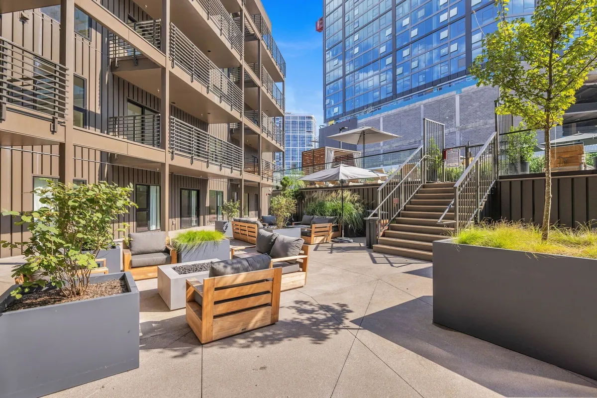 A contemporary outdoor seating area invites guests to relax or socialize, featuring modern furniture, planters, and stairs leading to another level. The setting is bright, open, and perfect for gatherings, meals al fresco, or enjoying seasonal events.