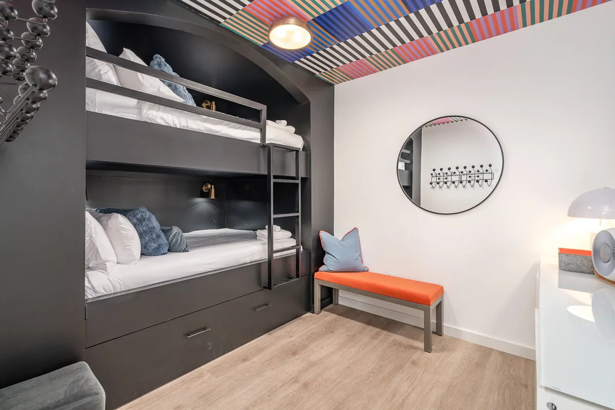 A fun and colorful bedroom designed with bunk beds, a playful ceiling, and a cheerful orange bench. The decor encourages creativity and a warm, friendly vibe, ideal for children or groups sharing a space.
