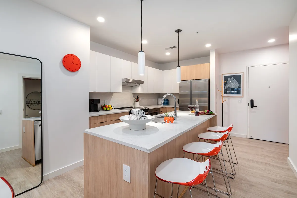 This image showcases a modern kitchen with a central island, stylish bar stools, a red accent clock, and up-to-date appliances. The space is bright and inviting, ideal for cooking, entertaining, or gathering around for a Southern-inspired brunch. The open layout visually connects this kitchen to adjacent living and dining spaces, reinforcing the open-concept feel.