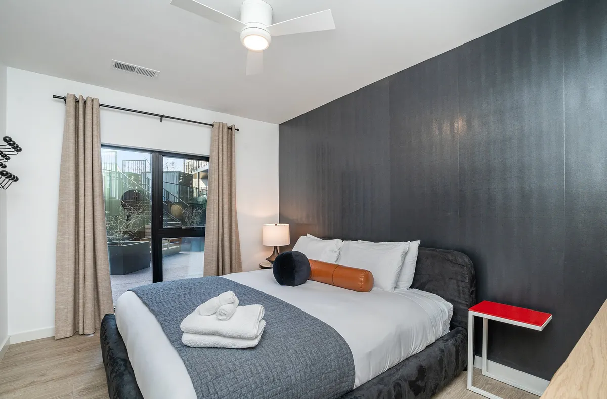 This image showcases a serene bedroom with a plush bed positioned near a window flooding the space with natural light. A nightstand with a lamp adds to the cozy, tranquil atmosphere, and the setup is ideal for peaceful relaxation, reading, or unwinding after a long day.