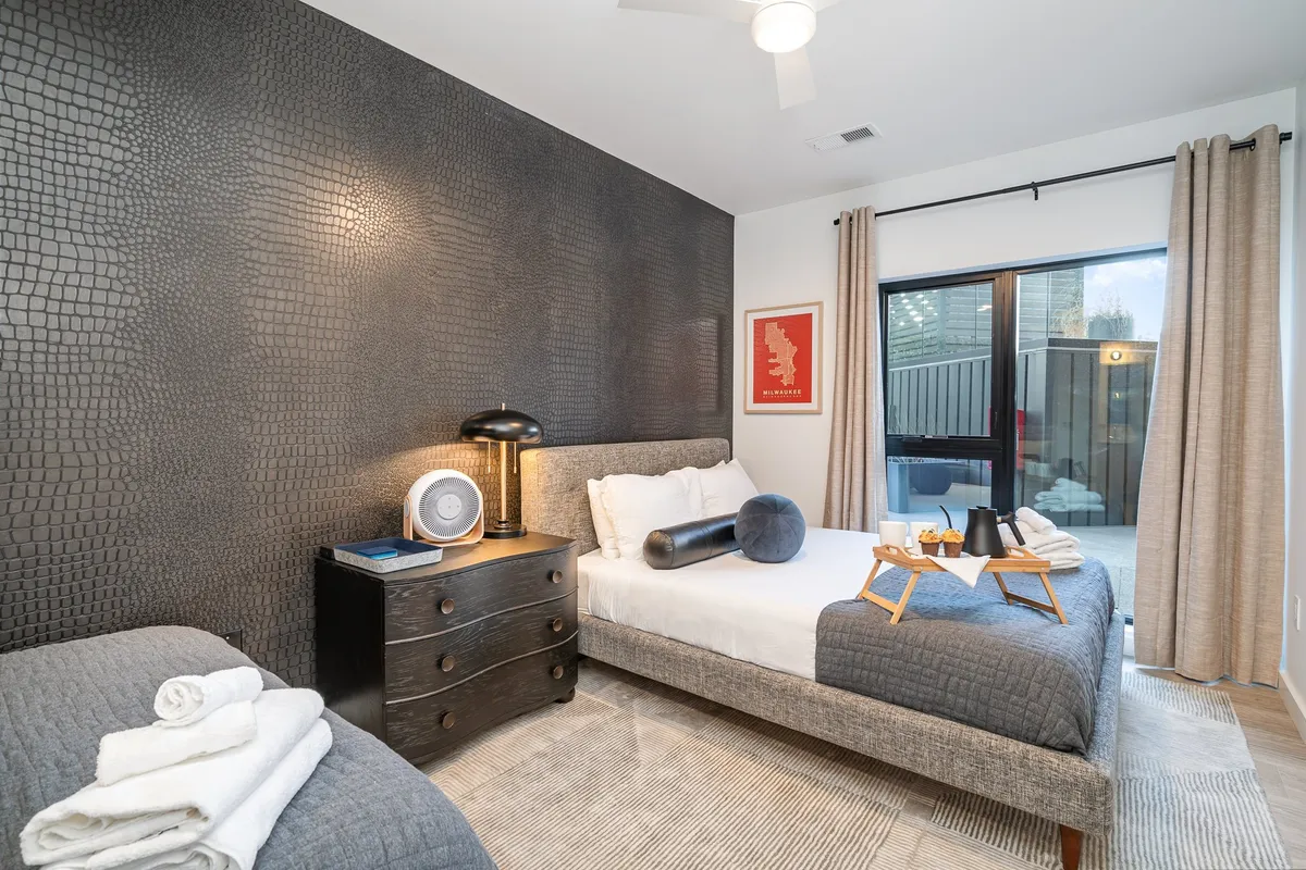 A serene bedroom with a textured black accent wall, a comfortable bed with white linens, a nightstand with a lamp and clock, and a window offering a city view. Warm lighting creates a tranquil ambiance above the bustling downtown.