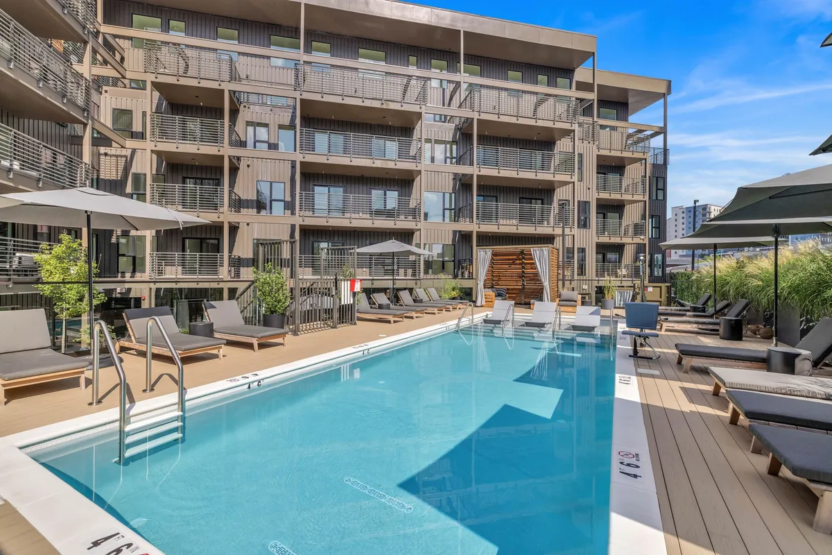 The image showcases a sparkling swimming pool set amidst sun loungers and outdoor umbrellas, with the building facade in the background. The scene is relaxed, with guests swimming, sunbathing, and socializing poolside, evoking a true resort atmosphere.